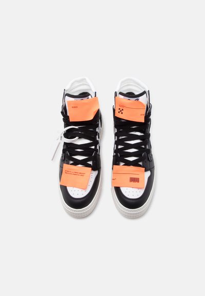 Ζευγάρι ασπρόμαυρα ψηλά sneakers με μαύρα κορδόνια, το καθένα καλυμμένο με τέσσερις φωτεινές πορτοκαλί ορθογώνιες ετικέτες με κείμενο και σύμβολα.