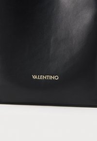Čierna kožená kabelka s hladkou textúrou a embosovaným zlatým logom "VALENTINO" na prednej strane. Minimalistický dizajn bez ďalších ozdôb.