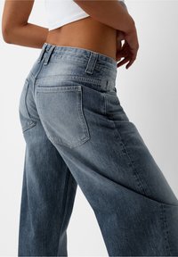 Person klädd i högmidjade, tvåtoniga blå jeans och en vit croppad topp, stående mot en ljus, enfärgad bakgrund.