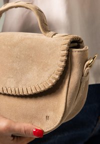 Beige suède handtas met een gebogen vorm, gestikte randen en een klein gouden hardware detail. Handvast met een gladde textuur.