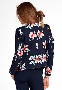 Kobieta z jasnobrązowymi włosami w luźnym kokie, ubrana w granatową marynarkę w kwiaty i dopasowane spodnie, zwrócona plecami w białym tle.