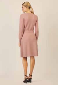 Knielanges, enges Kleid aus weichem rosa Strickstoff, mit langen Ärmeln, einer Taillenpartie und einem geraden Rock. Schwarze strappy High Heels.