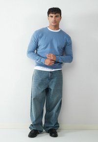 Jasnoniebieski sweter dziany na białej koszulce z okrągłym dekoltem, w połączeniu z luźnymi jasnoniebieskimi dżinsami i czarnymi butami, na białym tle.