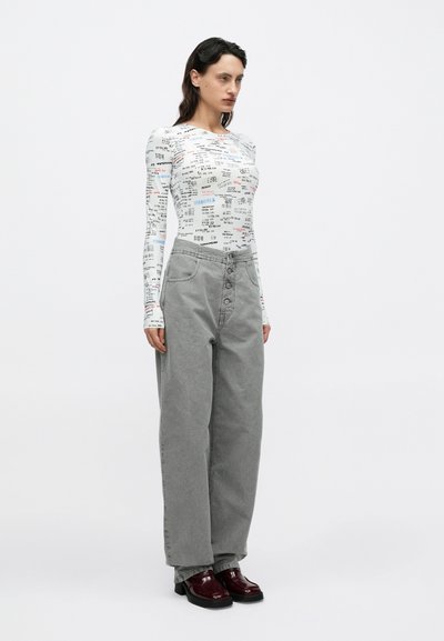 Biele tričko s dlhým rukávom a potlačou viacfarebných textov, v kombinácii s vysokým pásom, širokými sivými manšestráckymi nohavicami a lesklými bordovými topánkami.