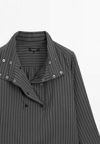 Chemise gris foncé à fines rayures avec un large col comportant plusieurs boutons-pression et une étiquette de marque visible à l'intérieur de l'encolure.