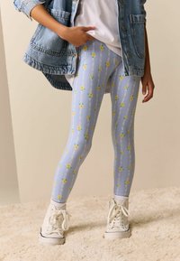 Modré legíny s květinovým vzorem s žlutými květy a zelenými listy, doplněné o džínovou bundu a bílé vysoké tenisky.