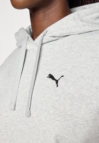 Siva hoodie izrađena od mekanog materijala s crnim vezenim logom. Uključuje vezice i detaljan prikaz područja oko vrata i pozicije loga.