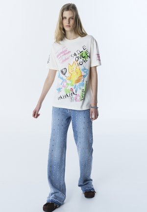 Giovane che indossa una maglietta bianca con schizzi colorati di gatti e fiori, abbinata a jeans azzurri decorati con piccole borchie.