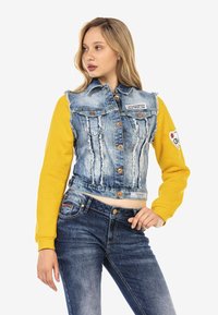 Veste en denim courte avec des manches longues jaunes, des détails déchirés, un système de boutonnière et un patch sur la manche gauche. Associée à un jean bleu.
