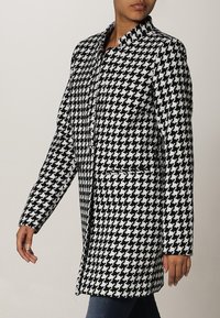 Svart och vit houndstooth-mönstrad kappa, medellång, med rak skärning, långa ärmar och två framfickor. Slät tygtextur.