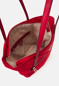 Czerwona torba na ramię z gładką powierzchnią, wyposażona w kieszenie na zamek błyskawiczny i kieszenie wsuwane wewnątrz, z wzorzystą beżową podszewką i zielonym logo.