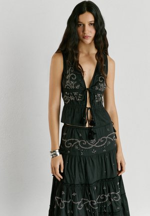 Mujer con cabello largo y oscuro, que lleva una blusa negra sin mangas bordada y una falda de capas a juego, de pie contra una pared clara y lisa.