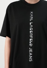 Asmuo vilki juodą marškinėlį su balta vertikaliais rašmenimis, kuriuose parašyta "KARL LAGERFELD JEANS" per patį vidurį.