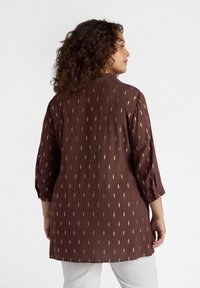 Blusa marrone con colletto, maniche a tre quarti e un sottile motivo diagonale dorato. Tessuto leggero con una finitura leggermente strutturata.