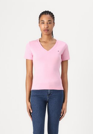 Femme aux cheveux tressés portant un t-shirt ajusté rose clair avec col en V et manches courtes, ainsi qu'un jean bleu foncé, debout devant un fond blanc.