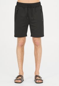 Svarta shorts med elastisk midjeband och dragsko, har två fickor med dragkedja på sidorna, knälånga, slät textur. Bärs med bruna sandaler.