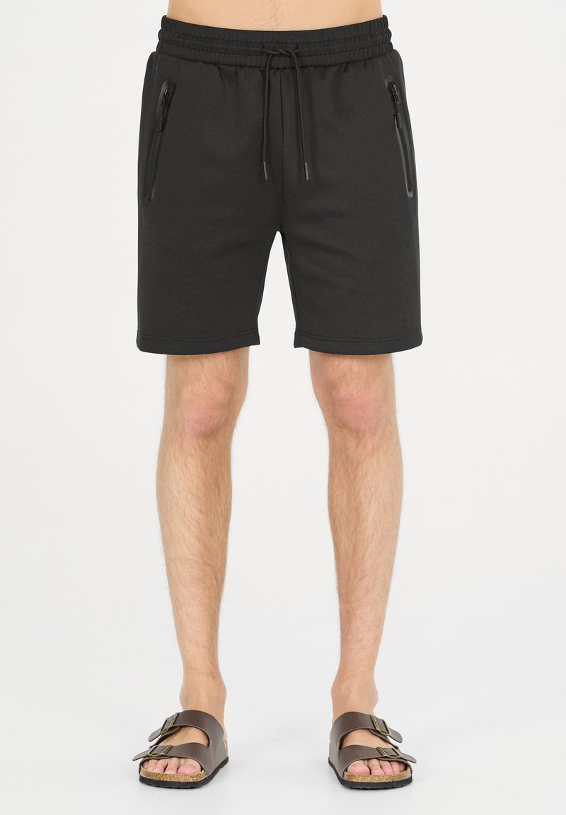 Svarta shorts med elastisk midjeband och dragsko, har två fickor med dragkedja på sidorna, knälånga, slät textur. Bärs med bruna sandaler.