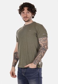 T-shirt verde oliva a maniche corte in morbido tessuto, con scollatura rotonda e un piccolo logo sul petto e sulla manica. Jeans in denim visibili.