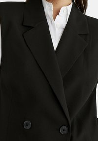 Schwarzer ärmelloser Blazer mit doppelter Knopfleiste, Reverskragen und zwei schwarzen Knöpfen. Glattes Material mit strukturierter Silhouette.