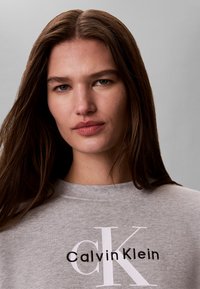 Νεαρή γυναίκα με μακριά καστανά μαλλιά που φοράει γκρι φούτερ Calvin Klein με απλό γκρι φόντο.