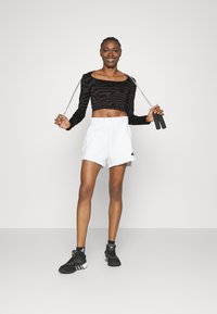Svart mønstrete langermet crop-top med rund hals, sammen med hvite shorts. Modellen holder et hoppetau og har på seg svarte treningssko.