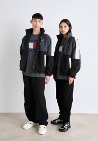 Dos modelos llevan chaquetas de color bloque a juego en gris, negro y crema, sobre camisetas grises con un logo. Las combinan con pantalones negros y zapatillas.