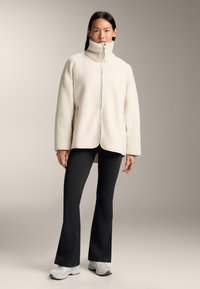 Giacca in pile color crema con colletto alto, chiusura con zip e tasche laterali, abbinata a pantaloni neri a zampa d'elefante e sneaker grigie chiare.