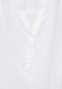 Tunique en coton blanc avec un col en V, dotée d'une patte de boutonnage avec six boutons et d'un détail de tissu froncé à l'arrière.