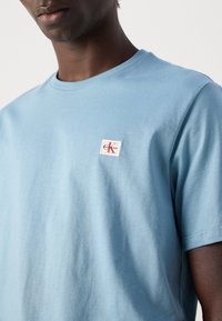 Moški nosi svetlo modro majico Calvin Klein z malim logotipom na levi strani na ozadju brez vzorca.
