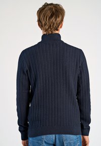 Dunkelblauer Strickpullover mit strukturiertem Rippmuster und hohem Kragen. Verfügt über lange Ärmel sowie ein eng anliegendes Bündchen und Saum.