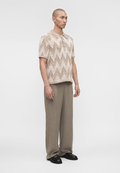 Άνδρας που στέκεται, φοράει πλεκτό πόλο με κοντά μανίκια και μπεζ ζιγκ ζαγκ μοτίβο, φαρδιά μπεζ παντελόνια και μαύρα παπούτσια σε απλό φόντο.