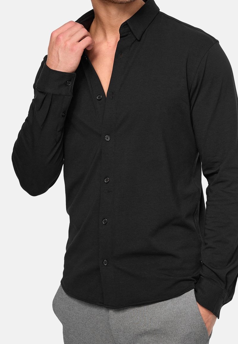 Homme ajustant le col d'une chemise noire à boutons, portant un pantalon gris, vue recadrée du torse et des bras.