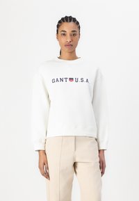 Sweatshirt blanc avec un col rond, présentant un texte brodé bleu marine "GANT U.S.A." et un emblème rouge et bleu. Tissu en coton, coupe décontractée.