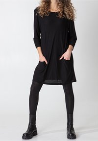 Svart långärmad klänning med rund halsringning, två frontfickor och avslappnad passform, kombinerad med svarta strumpbyxor och ankle boots.