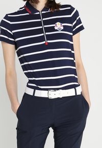 Polo bleu marine et blanc à rayures avec un zip court et un logo USA brodé, associé à un pantalon bleu marine et une ceinture blanche.