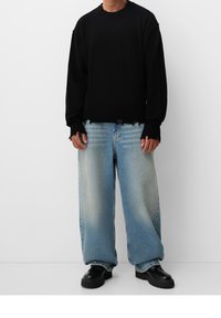 Schwarzer Strickpullover mit Rundhalsausschnitt und ausgefranzten Kanten, kombiniert mit hellblauen weiter geschnittenen Jeans und dickenschwarzen Schuhen.