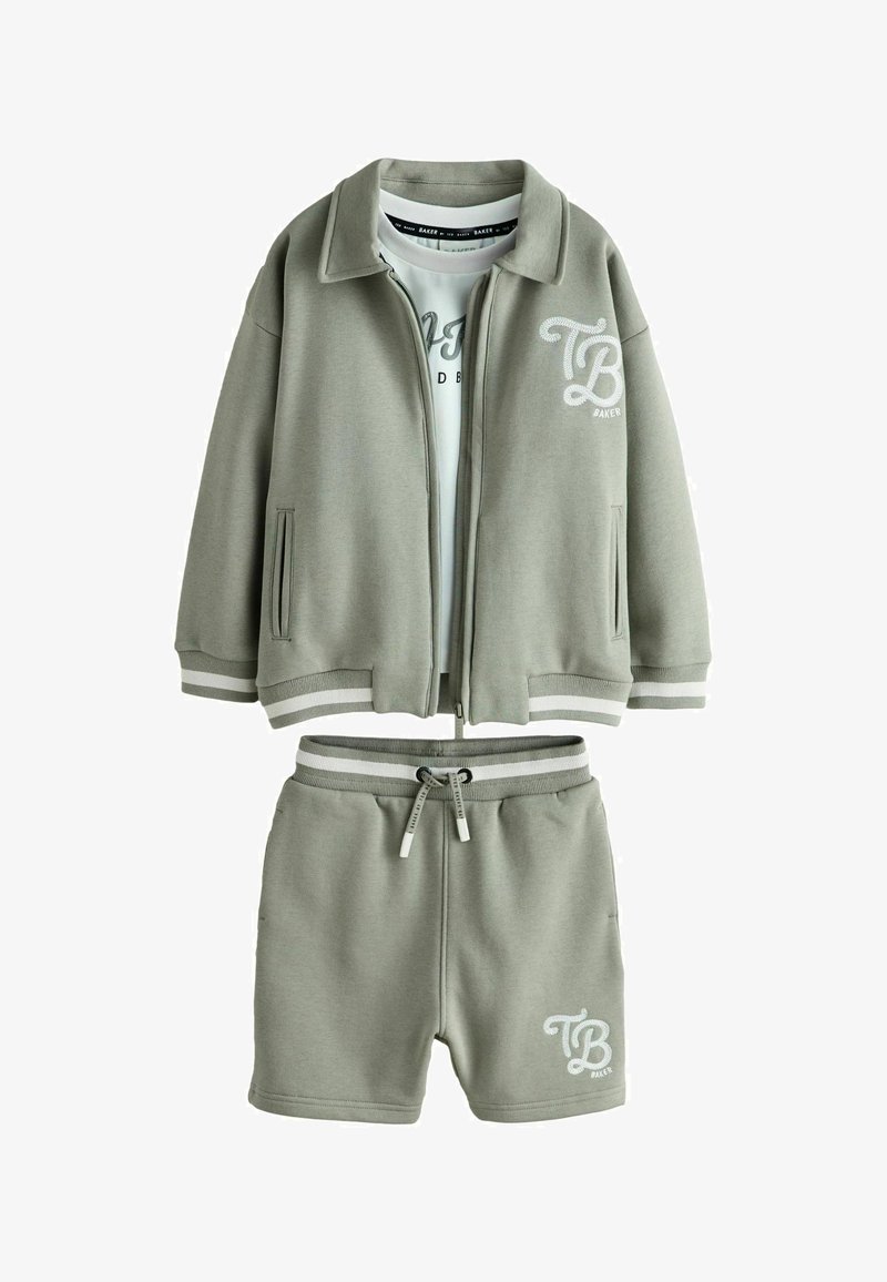 Jasnozielona kurtka na zamek i pasujące spodenki z białym logo „TB Baker” oraz paskowanymi mankietami, nałożone na białą koszulkę z grafiką.
