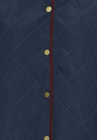 Mørkeblå quiltet jakke med diamantmønster, gullsnapknapper og kontrastfarget burgunderkant langs frontåpningen.