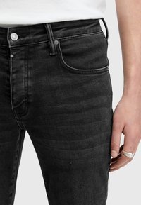 Černé džínové kalhoty s úzkým střihem, které mají pět kapes, jemné blednutí a viditelné švy. K tomu stříbrný prsten na ruce.