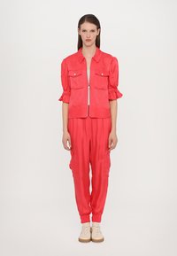 Aansluitend twee-delig outfit in felroze stof. Bevat een rits-up jack met korte mouwen en borstzakken en getailleerde broek met zijzakken.