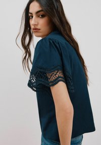 Marineblaues Shirt mit kurzen, gepufften Ärmeln, das an den Kanten mit aufwendiger Spitze verziert ist. Glatter Stoff mit lockerer Passform.