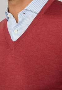 Niebiesko-biała w paski koszula z guzikami noszona pod czerwoną swetrem w serek na górnej części ciała osoby.