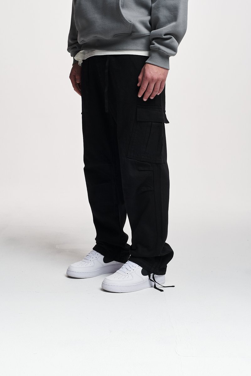 Black cargo pants with multiple pockets, relaxed fit, and drawstring waist. Worn with white sneakers and a gray hoodie above.