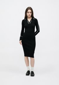 Zwart geribd jurkje met lange mouwen, V-hals en witte details. Gecombineerd met witte sokken en glanzende zwarte loafers.