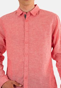 Chemise boutonnée corail clair avec un tissu texturé, col pointu et sept boutons sur le devant. Les manches longues sont retroussées aux poignets.