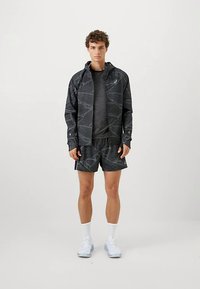Veste de sport noire à motif géométrique, associée à une chemise gris foncé et un short noir. Le mannequin porte des baskets bleu clair et des chaussettes blanches.