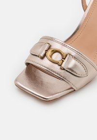 Metallic rose gold sandal with a square toe, a wide strap featuring a gold-tone circular buckle and two accent straps. Smooth texture.