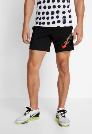 Mand iført sorte Nike shorts med rødt logo, hvide og sorte mesh løbesko med neongrøn sål og en hvid skjorte med prikker.