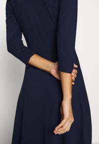 Personne portant une robe bleu marine à manches trois-quarts, bras plié derrière le dos tenant le poignet opposé sur un fond uni.