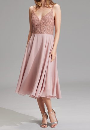 Frau trägt ein ärmelloses, staubig rosa Midikleid mit perlenverziertem Oberteil und fließendem Chiffonrock, kombiniert mit silbernen Absatzschuhen mit Knöchelriemen.